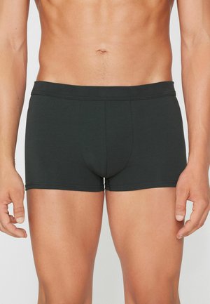 Torso masculino con calzoncillos bóxer negros ajustados, de pie con los brazos relajados a los lados sobre un fondo neutro.