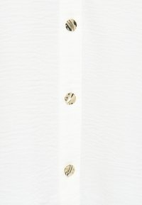 Άσπρο πουκάμισο με υφή, διακοσμημένο με μια σειρά από χρυσά κουμπιά με σχέδια που τρέχουν κατά μήκος της μπροστινής πλευράς.