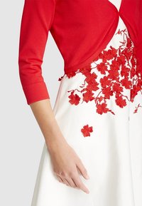 Rød cardigan med tre-kvartærmer over en hvid kjole, der har intrikate røde blomsterbroderier på nederdelen. Tekstureret og tætsiddende design.