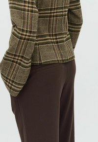 Persona che indossa una giacca a quadri marroni e pantaloni marroni scuri, fotografata dalla vita in giù con un angolo leggermente inclinato.