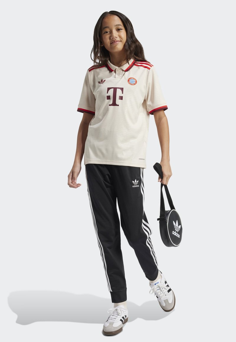 T-shirt de football beige clair avec des accents rouges, doté d'un col et de logos. Pantalons de jogging noirs avec des rayures blanches et une marque Adidas, sneakers blanches.