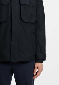 Water-resistant black jacket featuring two large flap pockets, a button closure, and adjustable cuff with contrasting inner lining.