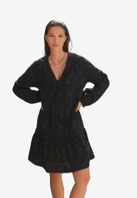 Robe noire à manches longues, décolleté en V profond, broderie florale, ourlet à volants et détails à cordon de serrage. Tissu léger, finition texturée.