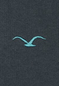 Grauer Stoff mit einem gestickten türkisfarbenen Möwen-Logo, mittelgroß und mit einem strukturierten Strickmuster.