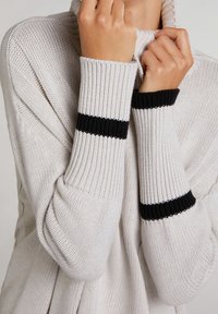 Maglione lavorato a maglia di un grigio chiaro con colletto alto e polsini a coste, caratterizzato da strisce nere. Il materiale è strutturato, morbido e con una vestibilità comoda.