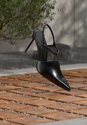 Scarpa nera con tacco alto a punta e cinturino al tallone, sospesa sopra un pavimento in assi di legno con vegetazione all'aperto sullo sfondo.