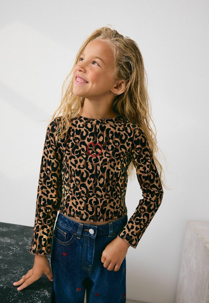 Jong meisje met lang, krullend blond haar draagt een top met luipaardprint met een rode hart en blauwe jeans met kleine rode borduursels, glimlachend en naar boven kijkend.