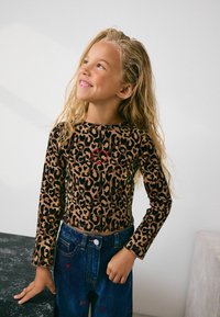 Noor tüdruk, kellel on pikad lokkis blondid juuksed, kannab leopardimustrilist topi punase südamega ja siniseid teksaseid, millel on väike punane tikand. Ta naeratab ja vaatab ülespoole.