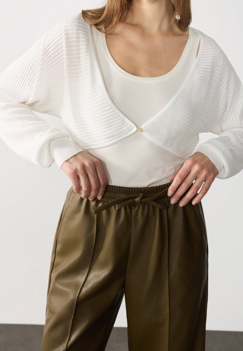 Vit, öppen stickad cardigan med knappstängning, kombinerad med en krämfärgad tanktop och olivgröna, veckade, högmidjade byxor i konstläder.