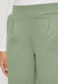 Un pantalon texturé vert avec une taille haute et des plis devant. Le tissu est lisse avec une légère élasticité pour plus de confort.