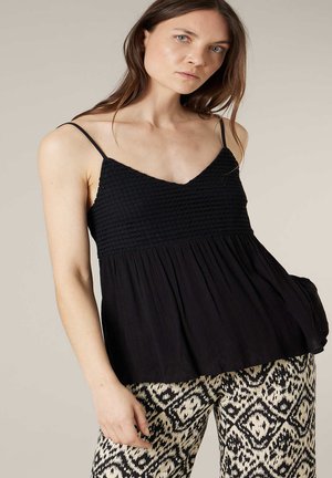 Vrouw draagt een zwart gestructureerd mouwloos topje met dunne bandjes en zwart-witte broek met patroon, poseert tegen een effen achtergrond.