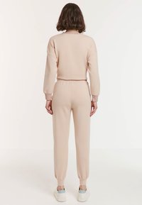 Beige trainingspak met een ronde hals en lange mouwen. Elastische tailleband en manchetten. Witte sneakers met lichtblauwe accenten. Achteraanzicht.