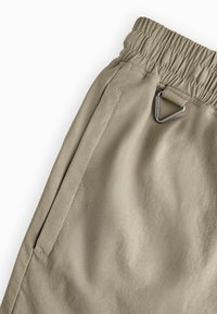 Beige broek met een elastische tailleband en een klein metalen lusdetail. De stof is glad, met een zijzak en gestikte naden.