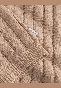 Beżowy sweter dziany w ściągacz, z pionowymi paskami i małą metką z logo "LES DEUX" na brzegu.