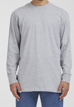Homme portant une chemise à manches longues gris clair unie et un jean bleu, debout devant un fond neutre avec les mains détendues le long du corps.