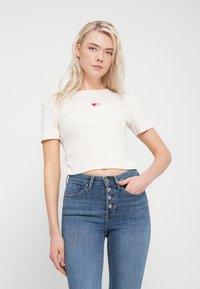 Μπλούζα τύπου crop top με κοντά μανίκια σε κρεμ χρώμα και ριμπ υφή, διακοσμημένη με δύο κόκκινες καρδιές κεντημένες, σε συνδυασμό με ψηλόμεσο μπλε τζιν παντελόνι με πέντε κουμπιά.