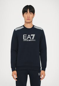 Felpa blu navy con logo bianco "EA7 EMPORIO ARMANI", polsini a costine e dettaglio a righe sulle spalle, realizzata in morbido tessuto testurizzato.