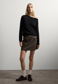 Maglione nero off-shoulder, mini gonna marrone con tasche frontali, calze bianche a coste e scarpe marroni scure, su uno sfondo neutro.