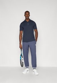 Camisola polo azul marinho com um logo discreto, combinada com calças de moletom cinzentas com logo e riscas brancas nas bainhas, usadas com sapatilhas brancas.