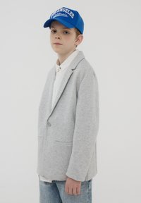 Blazer in cotone grigio chiaro con un solo bottone, abbinato a una camicia bianca e un cappellino da baseball blu con la scritta "Los Angeles California".