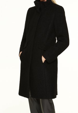 Kobieta nosząca czarny teksturowany płaszcz o średniej długości z wysokim kołnierzem i rękami w przednich kieszeniach, w połączeniu z czarnymi spodniami.