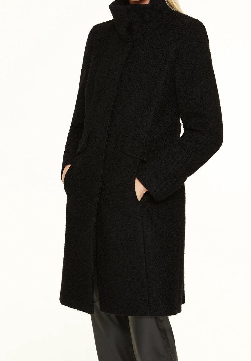 Femme portant un manteau texturé noir mi-long avec un col montant et les mains dans les poches avant, assorti à un pantalon noir.
