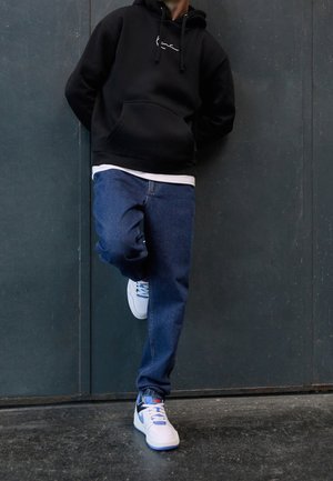 Felpa con cappuccio nera con tasca frontale, abbinata a jeans blu e sneakers bianche con dettagli blu. Appoggiato a un muro.