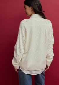 Pull crème avec un motif en losanges, orné de petites clous sur les épaules, présentant une coupe décontractée et un ourlet côtelé.