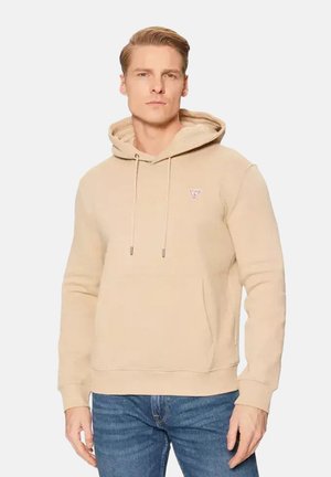 Jonge man met kort blond haar die een beige capuchontrui en blauwe spijkerbroek draagt tegen een effen lichte achtergrond.
