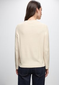 Top crema a maniche lunghe con finitura testurizzata, scollo rotondo e vestibilità rilassata, abbinato a jeans blu scuro con tasche posteriori.