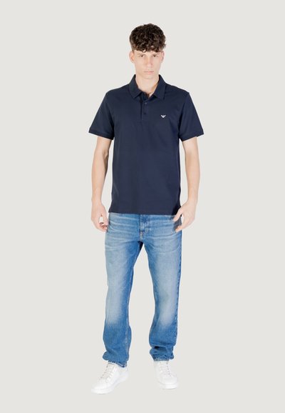 Mornarska polo majica s kratkimi rokavi, z dvojnim gumbom in vezenim logotipom. Usklajena z svetlo modrimi jeans hlačami in belimi supergami.