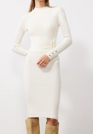 Kobieta w dopasowanej, białej, prążkowanej sukience z długimi rękawami i guzikami przy mankietach, w połączeniu z beżowymi zamszowymi kozakami za kolano.