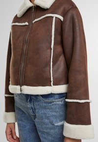 Giacca corta in similpelle marrone con colletto e rifiniture in pelliccia sintetica bianca, dotata di tasche frontali e chiusura con zip.