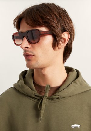 Braune rechteckige Sonnenbrille mit Verlaufsgläsern und dickem Rahmen, getragen zu einem grünen Kapuzenpullover mit Kordelzug und kleinem Logo.