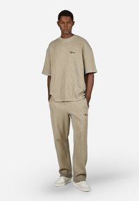 Mężczyzna w beżowej, prążkowanej koszulce z krótkim rękawem i pasujących spodniach z małym czarnym logo, stoi z rękami w kieszeniach i białych sneakersach.