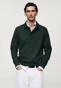 Dark green pullover with a collar, three-button placket, and ribbed cuffs. Worn over a white collared shirt. Styled with white pants.