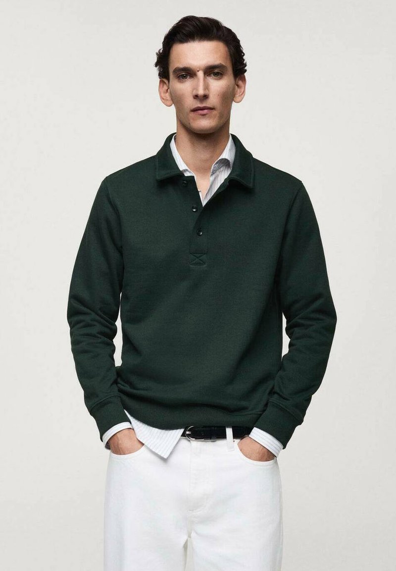 Dark green pullover with a collar, three-button placket, and ribbed cuffs. Worn over a white collared shirt. Styled with white pants.