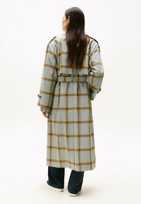 Abrigo tipo gabardina de cuadros azul claro con acentos en amarillo y marrón, cinturón en la cintura y mangas anchas. Se lleva sobre pantalones oscuros y zapatos blancos.