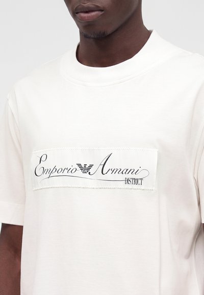 Άνδρας που φοράει λευκό T-shirt με ορθογώνιο πατρόν που φέρει το λογότυπο Emporio Armani και κείμενο σε μαύρο σενάριο.