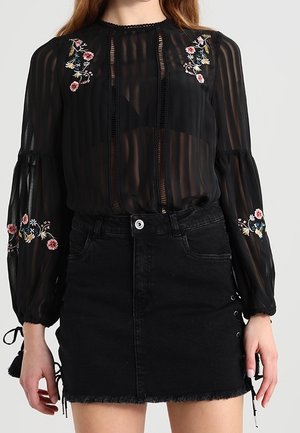 Femme portant un chemisier noir transparent avec broderies florales et une jupe en jean noire avec des côtés lacés et un ourlet effiloché.