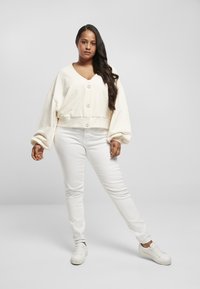 Geknipte crèmekleurige sweatshirt met oversized mouwen en knopen aan de voorkant, gecombineerd met witte skinny jeans en witte sneakers, tegen een lichtgrijze achtergrond.