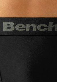 Juodas elastinis diržas su šviesiai pilku įspaudu „Bench“, pritvirtintas prie juodo audinio gaminio, demonstruojančio lygią tekstūrą.