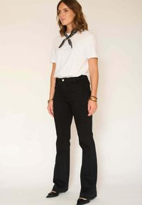 Pantalon en denim noir évasé à taille haute, associé à un t-shirt blanc et une écharpe noire, présentant un design épuré et un équipement minimal.