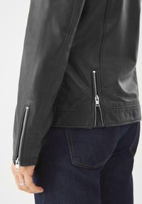 Schwarze Lederjacke mit silbernen Reißverschlüssen an den Ärmeln und Seiten, mit glatter Textur und taillierter Passform. Kombiniert mit dunkelblauen Jeans.