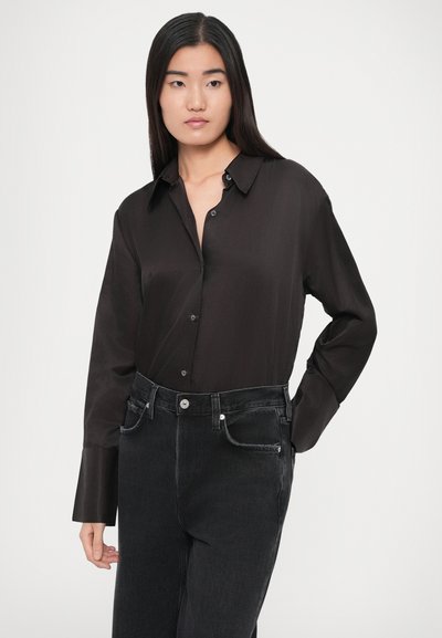 Μαύρο σατέν πουκάμισο με μακριά μανίκια που διαθέτει γιακά και κουμπιά, συνδυασμένο με μαύρα τζιν ψηλόμεσα. Λείο ύφασμα και εφαρμοστή σιλουέτα.