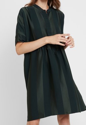 Woman wearing loose, short-sleeve, knee-length dress with wide vertical dark green stripes and a round neckline with buttons.