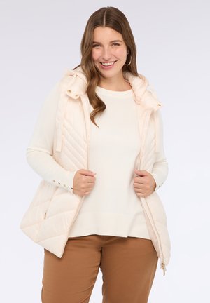 Lachende vrouw met lang haar in een crèmekleurige gewatteerde vest over een witte trui en een broek in bruin, terwijl ze het vest openhoudt.