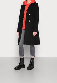 Zwarte wollen jas met dubbele knopen, gedragen over een rode hoodie. Grijze jeans en zwarte enkellaarsjes maken de outfit compleet. Over het geheel is er een gladde textuur.