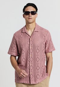 Kurzärmliges, pinkes, offenes Strickshirt mit Diamantmuster, ausgestattet mit einer Knopfleiste und Seitentaschen. Trägt eine dunkle Sonnenbrille.
