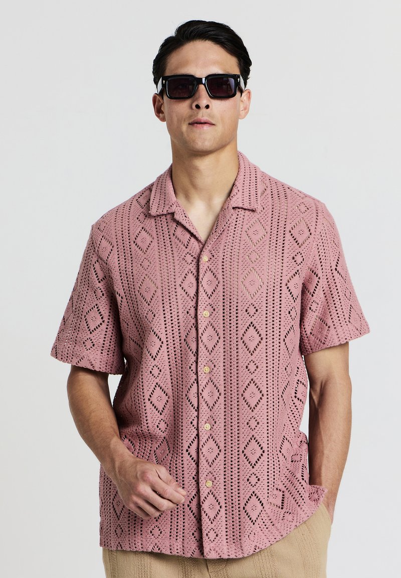 Kurzärmliges, pinkes, offenes Strickshirt mit Diamantmuster, ausgestattet mit einer Knopfleiste und Seitentaschen. Trägt eine dunkle Sonnenbrille.